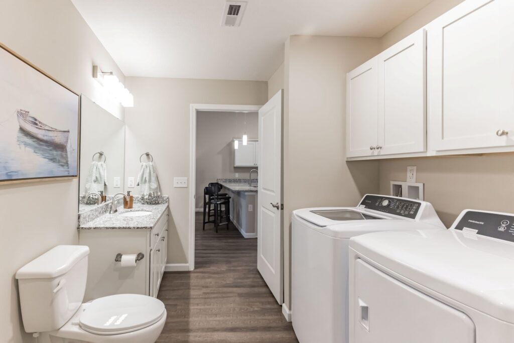 Modern laundry room with white cabinets, a washing machine, dryer, granite countertop, toilet, and wall art. The space features wood flooring and neutral-colored walls.