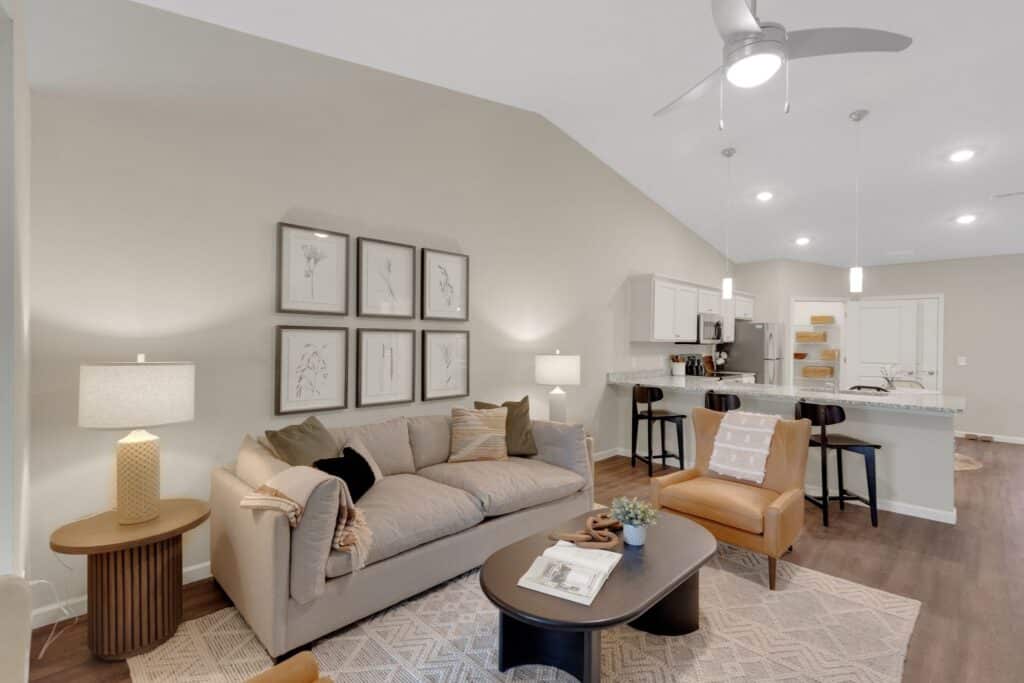 Modern living room with a beige sofa, two chairs, a coffee table, and framed botanical prints on the wall. The space is open to a kitchen with a bar counter and barstools.