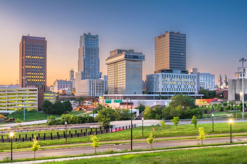 Downtown cityscape with modern high-rise buildings, green park in the foreground, and a clear sky at sunset.