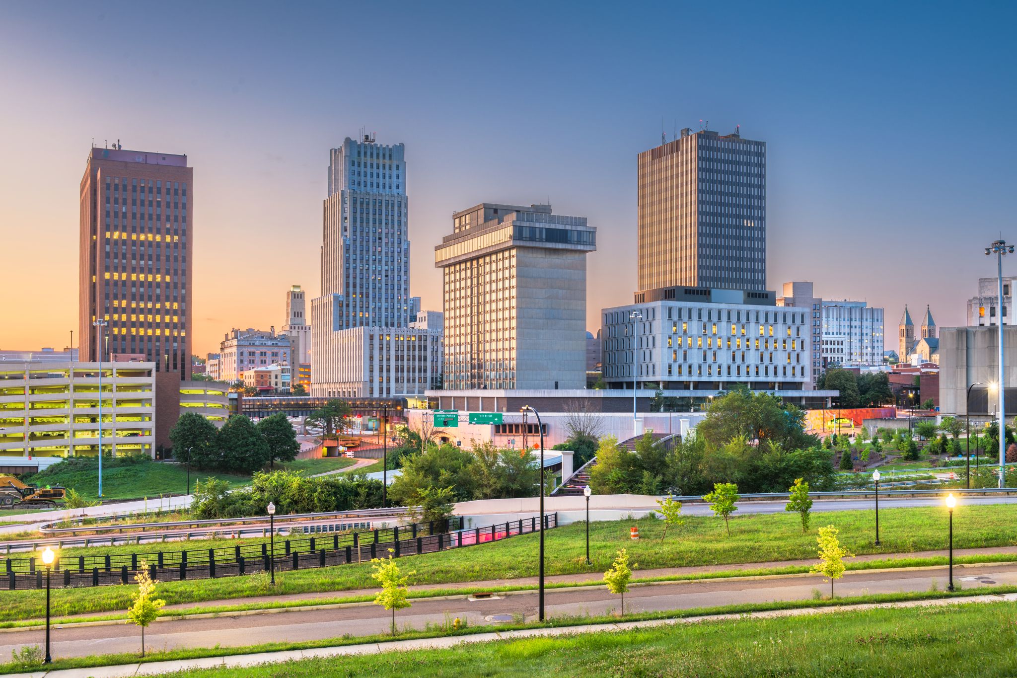 Downtown cityscape with modern high-rise buildings, green park in the foreground, and a clear sky at sunset.