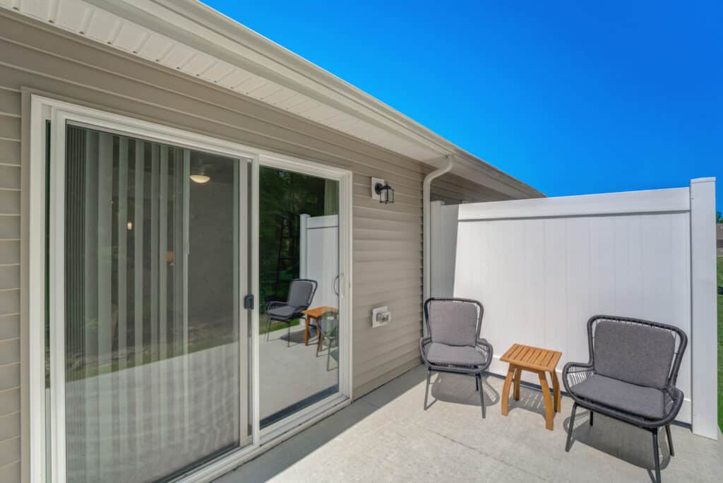 Small patio with two gray cushioned chairs and a wooden side table next to a sliding glass door and privacy fence on a sunny day.
