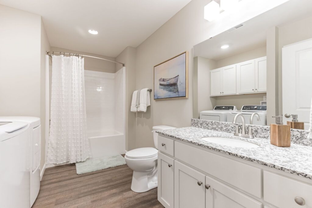 Bright bathroom with white cabinets, granite countertop, toilet, bathtub with shower curtain, washer and dryer, towel rack, and boat artwork on the wall.