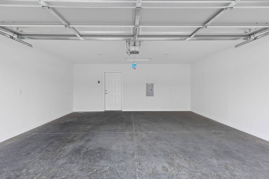 Empty two-car garage with concrete floor, white walls, ceiling-mounted garage door opener, and a single door on the left wall.