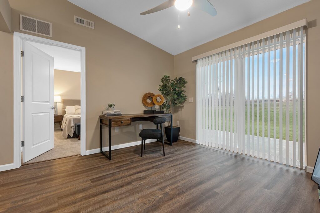 Room with wood flooring, a small desk and chair, potted plant, wall art, and large sliding glass door with vertical blinds overlooking a grassy yard. Bedroom visible through open door.