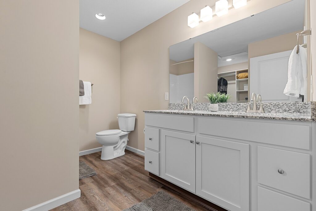 Modern bathroom with a double-sink vanity, granite countertop, large mirror, toilet, towel rack, and wood-look flooring under bright lighting.