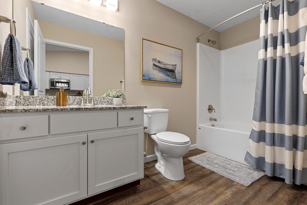 Bathroom with white cabinets, granite countertop, toilet, tub with striped shower curtain, wall art of a boat, and wood-look flooring. Towels hang on the wall and a rug is by the tub.