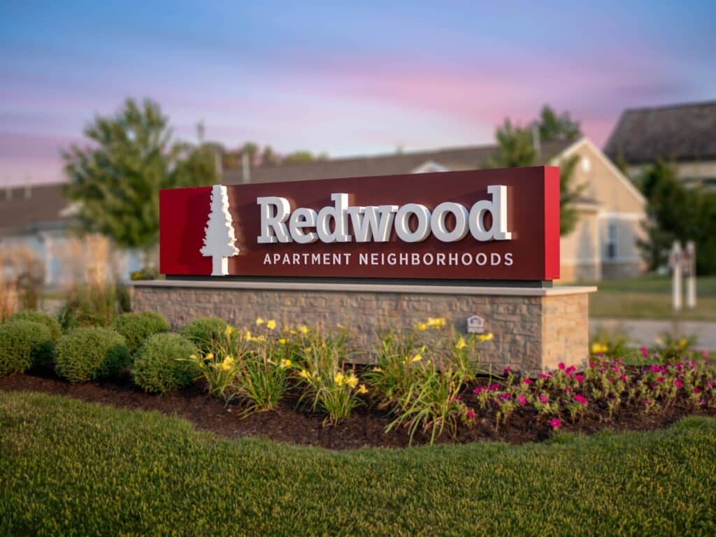 A sign reading “Redwood Apartment Neighborhoods” stands in landscaped grass with flowers and bushes, buildings visible in the background at sunset.