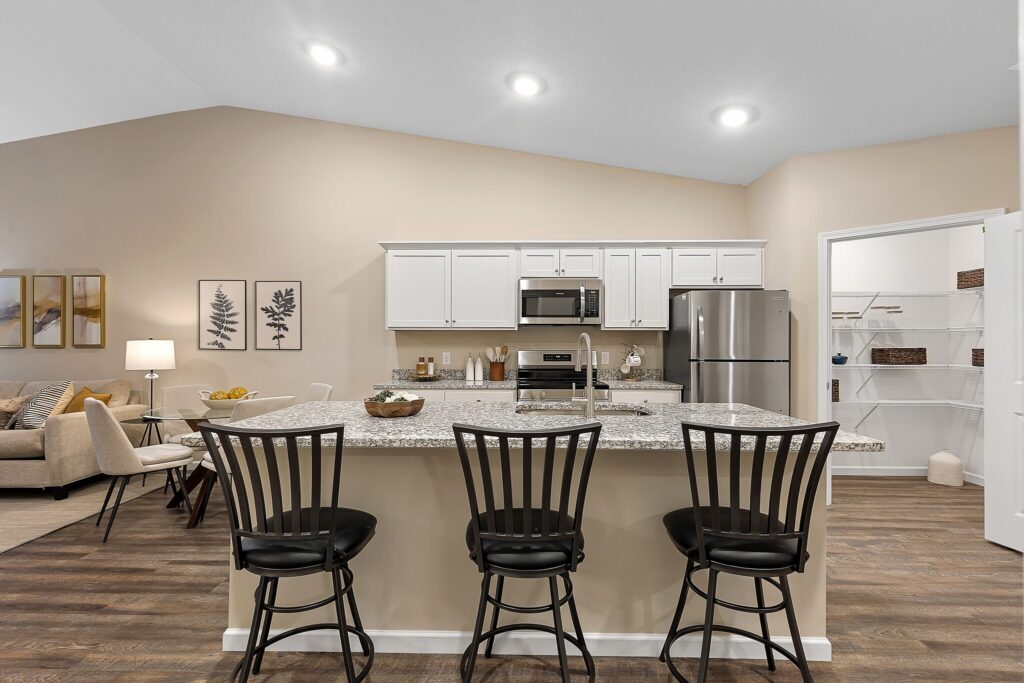 Modern kitchen with white cabinets, granite island, three black barstools, stainless steel appliances, and open pantry; living area visible in the background.