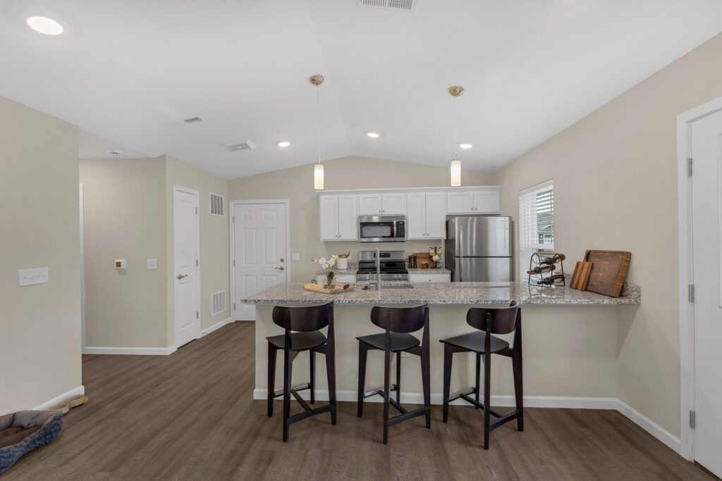 Modern kitchen with white cabinets, stainless steel appliances, granite countertops, and three black bar stools at a breakfast bar. Wooden floors and neutral walls complete the space.