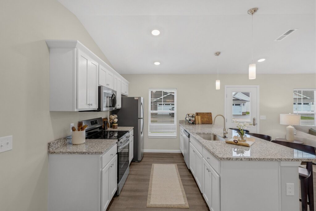 Modern kitchen with white cabinets, stainless steel appliances, granite countertops, pendant lighting, and a central island with a sink overlooking an open living area and large windows.