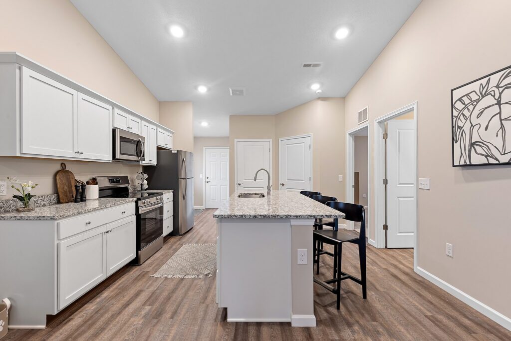 Modern kitchen with white cabinets, stainless steel appliances, a granite island with sink and seating, wood flooring, and neutral-colored walls.