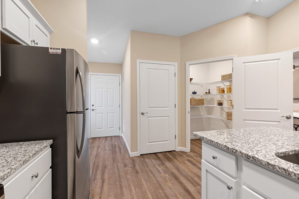 Modern kitchen with granite countertops, stainless steel refrigerator, wood flooring, white cabinets, and a walk-in pantry with shelves and storage baskets. Multiple white doors lead to other rooms.