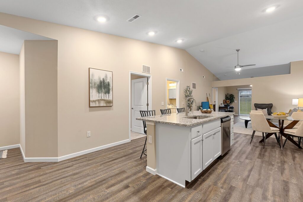Open-concept kitchen with an island, barstools, granite countertops, and wood flooring, extending into a dining and living area with neutral-toned walls and modern lighting.