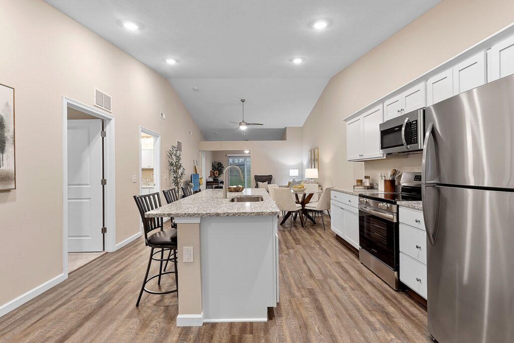 Modern open-concept kitchen with white cabinets, stainless steel appliances, a center island with bar stools, and wood flooring, leading to a living area with a ceiling fan.