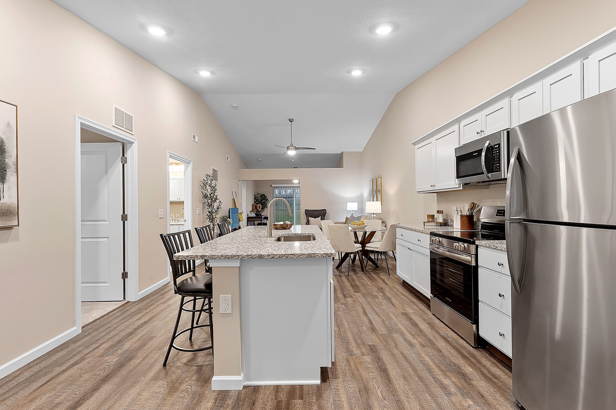 Modern open-concept kitchen with white cabinets, stainless steel appliances, a center island with bar stools, and wood flooring, leading to a living area with a ceiling fan.