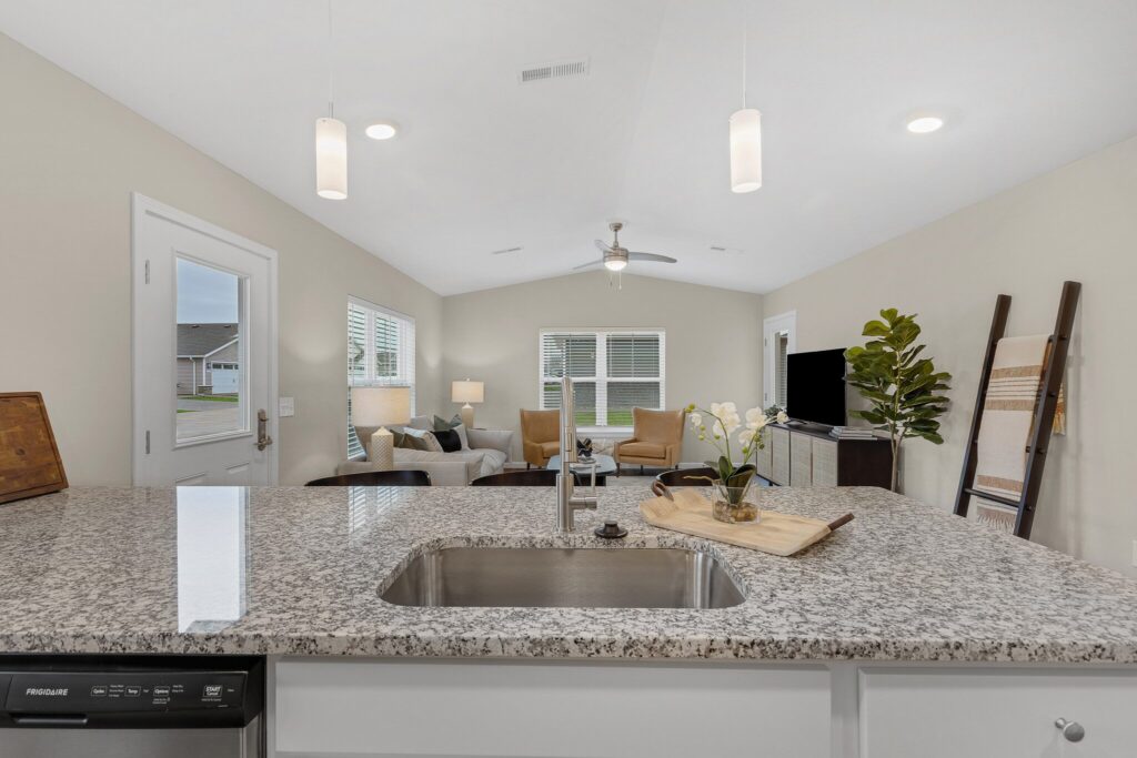 View from a kitchen island with a sink overlooking a living room with armchairs, TV, large window, plant, and decorative ladder.