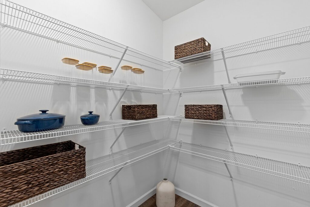 Empty wire shelving in a pantry with a few woven baskets, ceramic containers, blue cookware, a white dish, and a large jug on the floor.