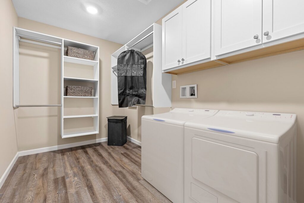 Laundry room with washer, dryer, white cabinets, open shelving with baskets, hanging rods, a garment bag, and a trash bin on wood-style flooring.