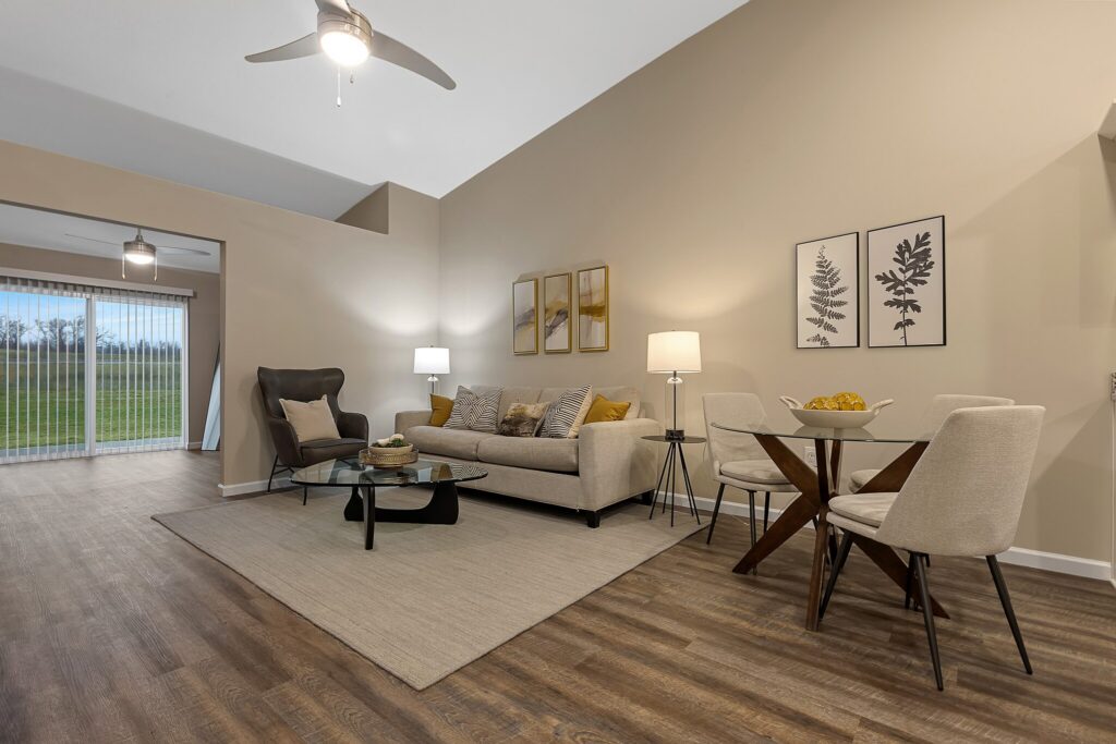 Modern living and dining area with beige walls, wood flooring, a sofa, armchair, coffee table, round dining table, and four chairs. Large window and sliding door in the background.
