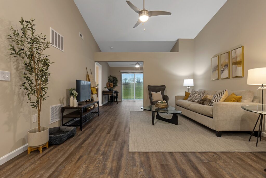 Modern living room with a beige sofa, glass coffee table, TV stand, potted plant, and decor. Wood flooring, neutral walls, ceiling fan, and sliding glass door with outdoor view.