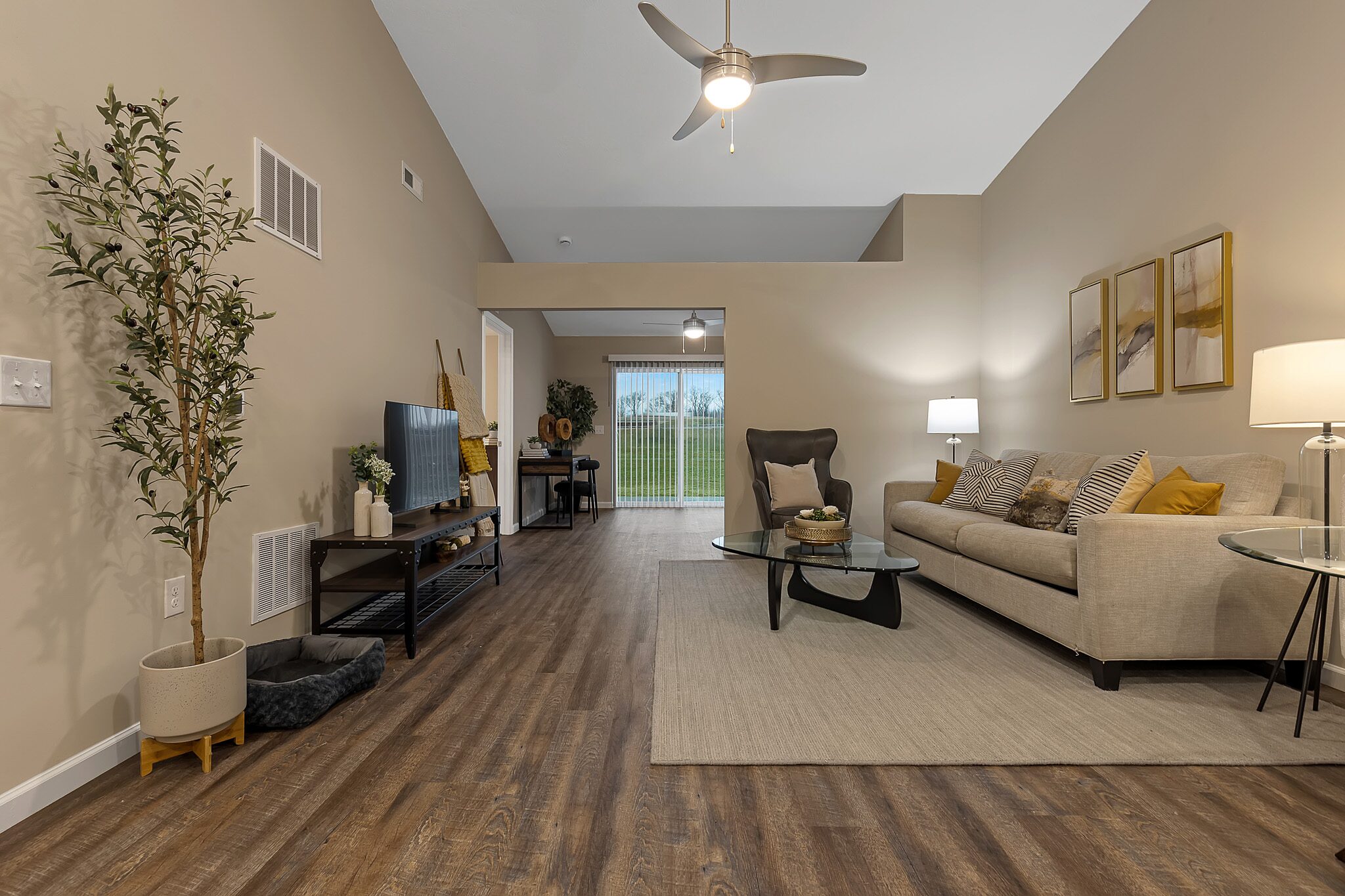 Modern living room with a beige sofa, glass coffee table, TV stand, potted plant, and decor. Wood flooring, neutral walls, ceiling fan, and sliding glass door with outdoor view.