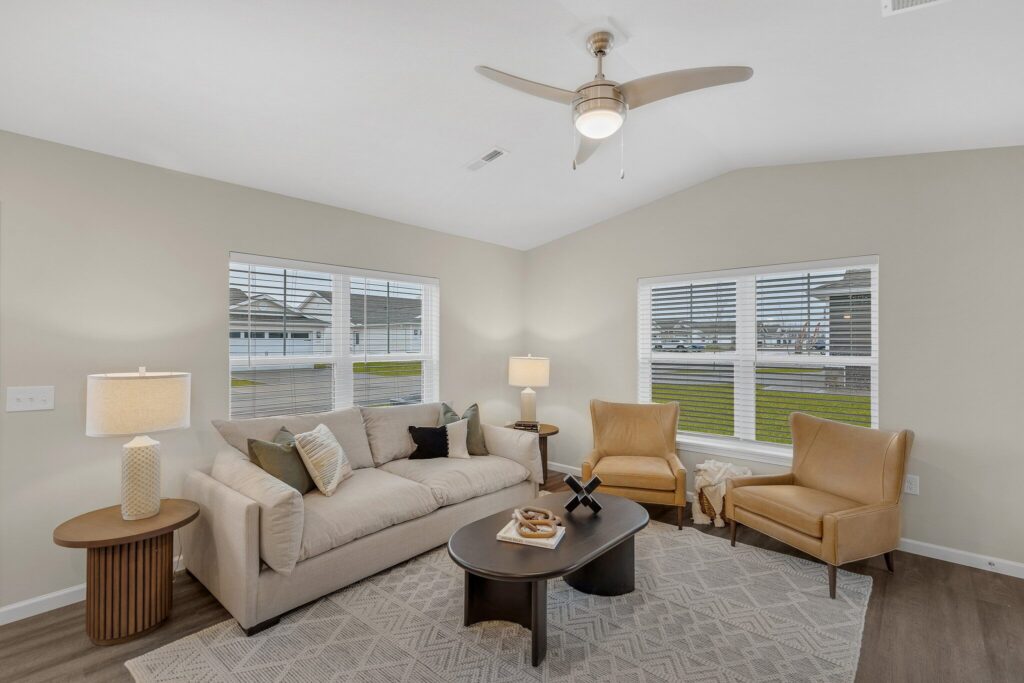 Bright living room with beige walls, a sofa, two tan armchairs, coffee table, lamps, and large windows with blinds, featuring modern decor and neutral colors.