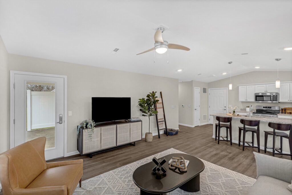 Modern open-concept living room and kitchen with neutral tones, a TV on a cabinet, armchair, indoor plant, and breakfast bar with three stools. Ceiling fan and recessed lighting overhead.