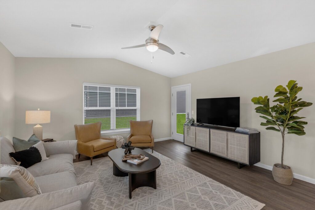 Neutral living room with beige walls, two tan chairs, gray sofa, coffee table, TV on a stand, potted plant, and a ceiling fan. Large window and glass door let in natural light.