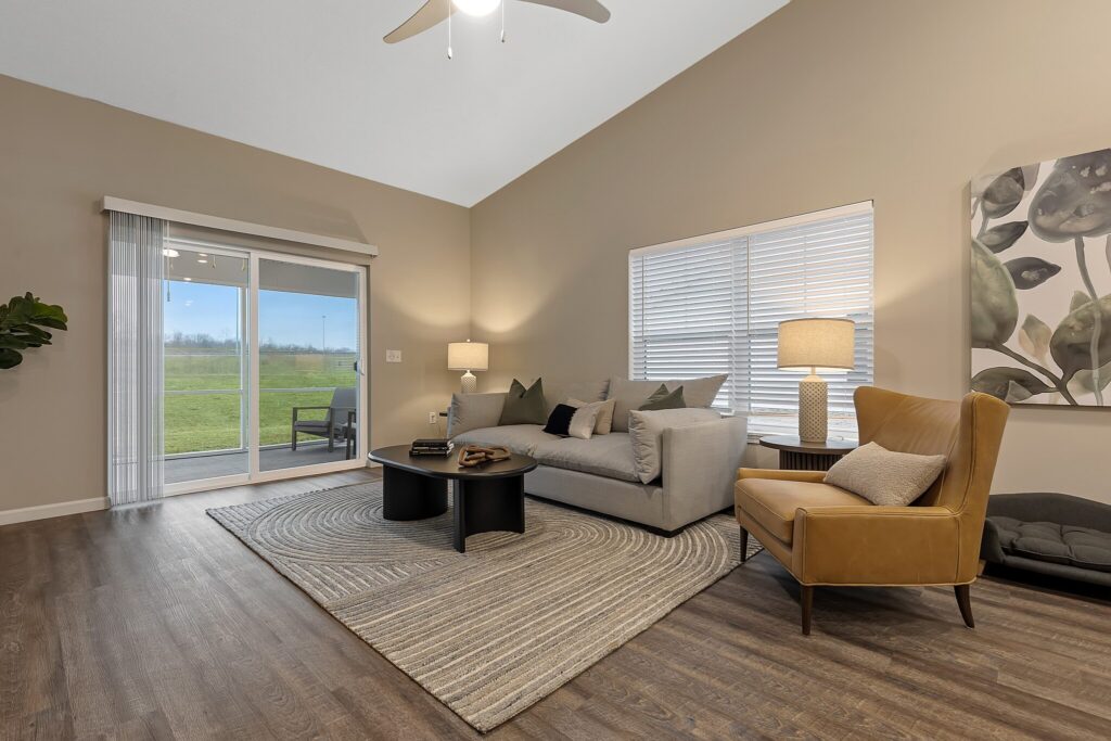 Modern living room with a gray sofa, tan armchair, round coffee tables on a striped rug, lamps, and large windows overlooking a grassy yard.