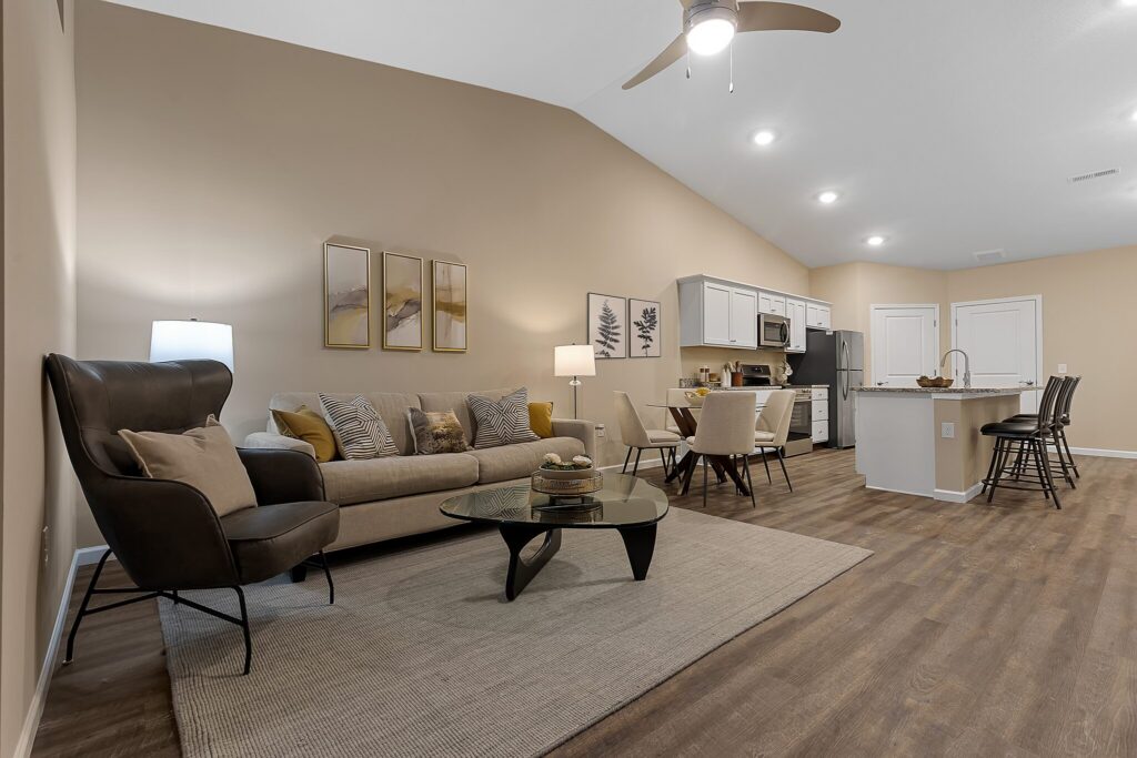 Modern open-concept living room and kitchen with neutral decor, gray sofa, armchair, glass coffee table, and bar stools at island on wood-look flooring under vaulted ceiling with ceiling fan.