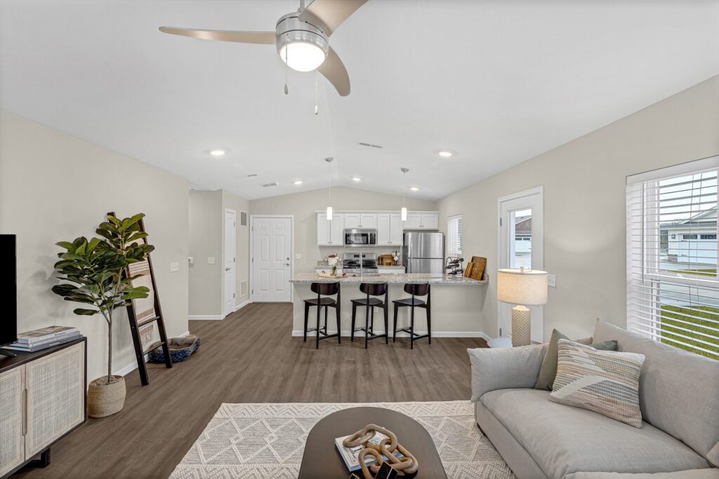 Open living room and kitchen with modern decor, bar seating at kitchen island, light walls, wooden floor, ceiling fan, and large windows letting in daylight.