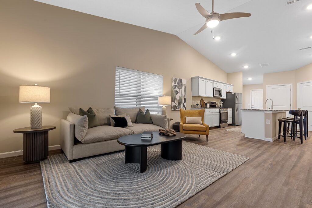 Modern open-concept living room and kitchen with a beige sofa, yellow armchair, round coffee tables, neutral decor, wood flooring, and a ceiling fan with lights.
