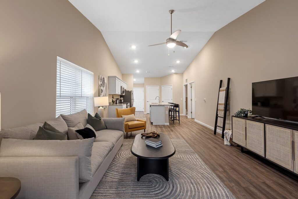 Modern living room with beige walls, gray sofa, yellow armchair, TV on cabinet, coffee table, and kitchen visible in the background with barstools and pendant lights.