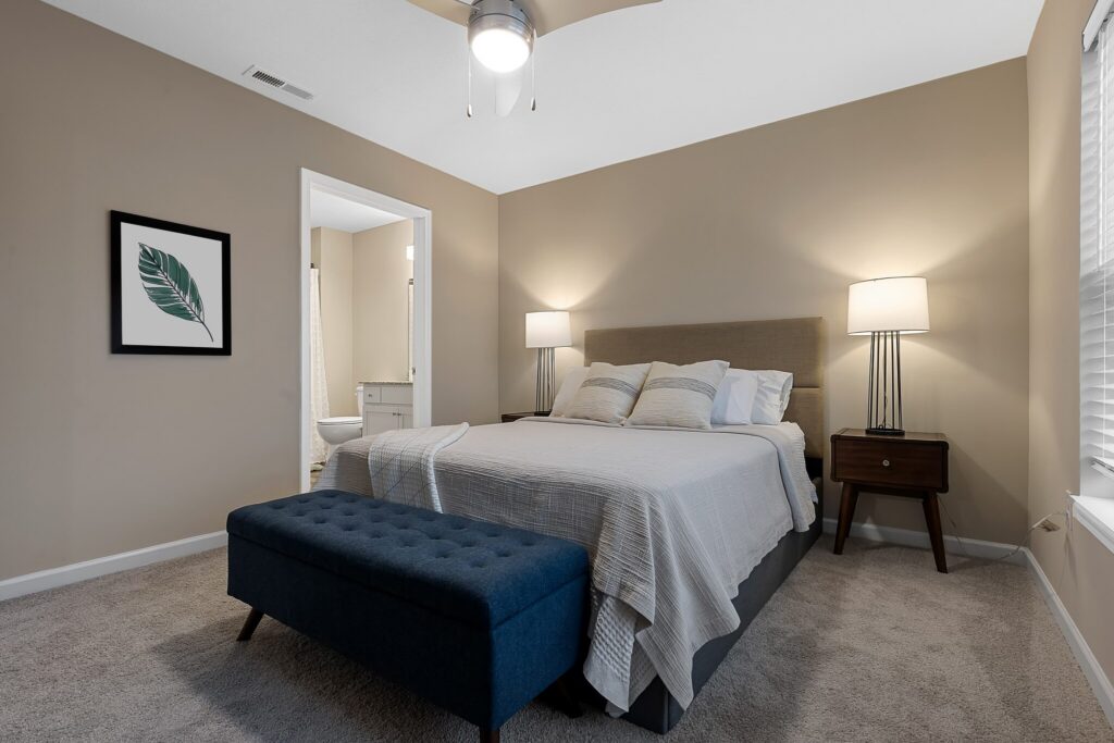Modern bedroom with beige walls, a bed with light bedding, a blue bench at the foot, two lamps, a nightstand, and a framed leaf print; door opens to a bathroom.