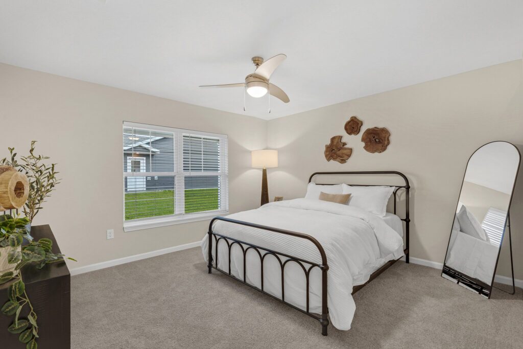 Bedroom with beige walls, a double bed with white bedding, a ceiling fan, a standing mirror, a lamp, wall decor, a dresser with plants, and a window showing a grassy yard.