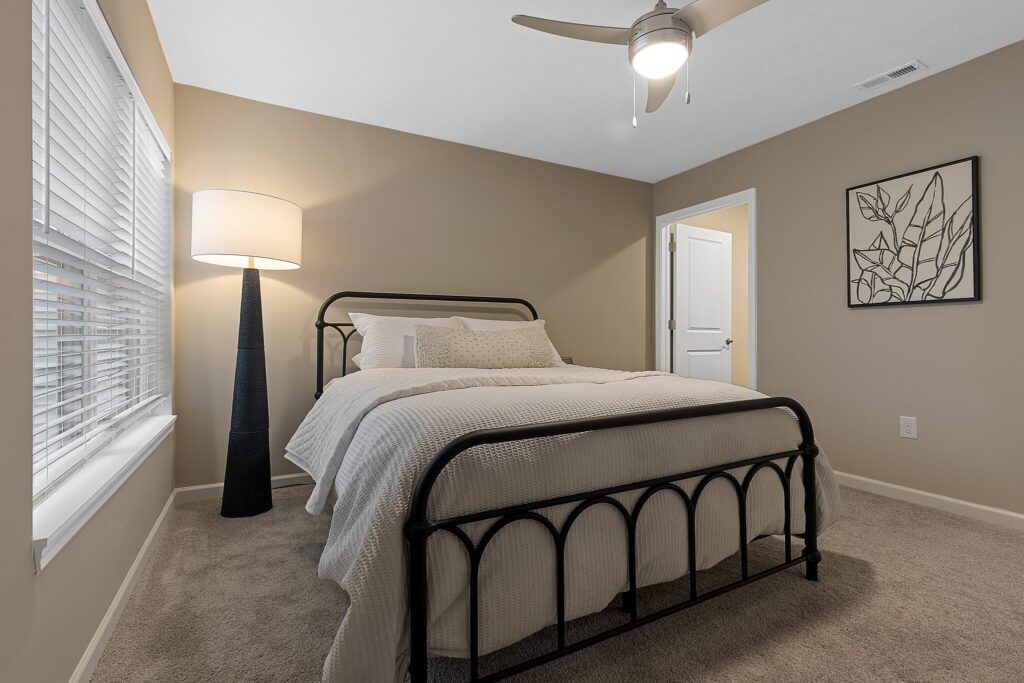 Bedroom with a black metal bed frame, white bedding, beige walls, a tall floor lamp, a window with blinds, and a framed abstract line art on the wall.