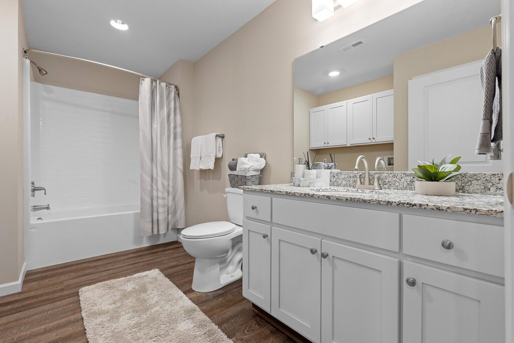 Modern bathroom with white cabinets, granite countertop, single sink, white tub with shower and curtain, wood-look flooring, and a beige wall. Towels and toiletries are neatly arranged.