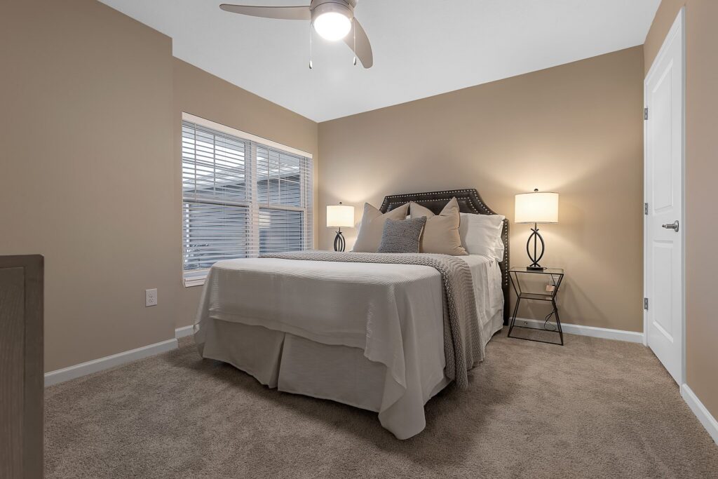 A neatly made bed with neutral bedding, two bedside tables with lamps, beige walls, carpeted floor, window with blinds, and a ceiling fan in a tidy bedroom.