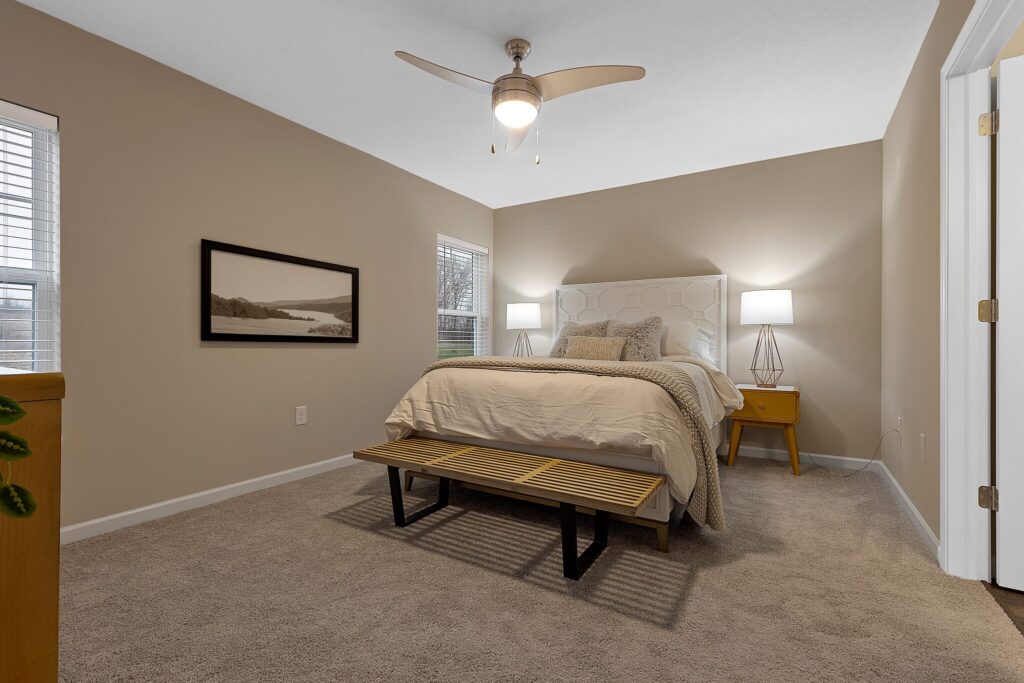 Bedroom with beige walls, carpeted floor, a bed with white bedding, two bedside tables with lamps, a ceiling fan, a wooden bench at the bed’s foot, and two windows.