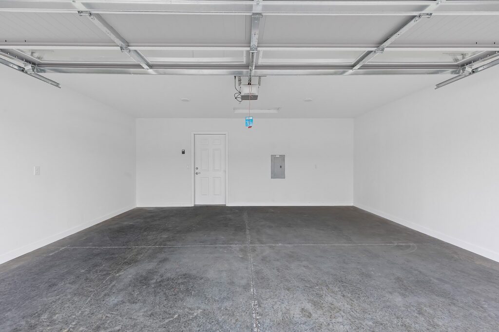 Empty two-car garage with a concrete floor, white walls, ceiling-mounted garage door opener, and a closed white door at the back.