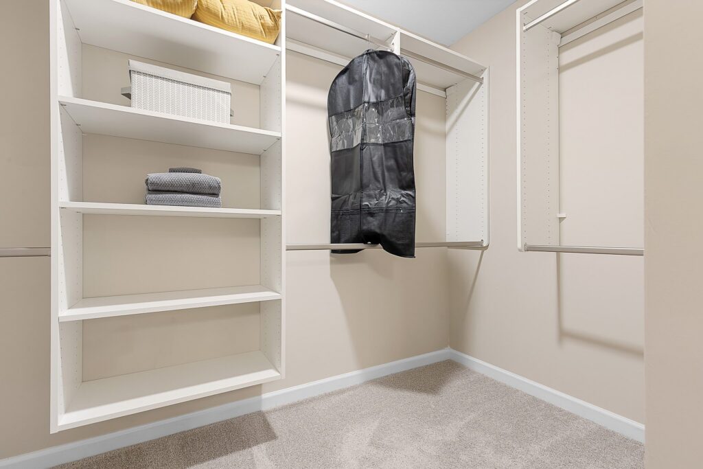 Empty walk-in closet with beige walls, white shelves holding towels and a box, and a black garment bag hanging on a rod.